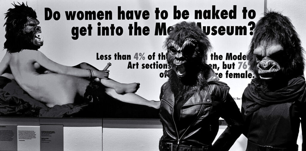 Black and white photograph of two of the Guerrilla Girls (https://en.wikipedia.org/wiki/Guerrilla_Girls) in front of a photo of the painting "The Grand Odalisque" by Ingres (https://en.wikipedia.org/wiki/Grande_Odalisque). The two women wear gorilla masks and a gorilla mask has been pasted onto the figure in the painting. Headline: "Do women have to be naked to get into the Met Museum?" Subhead: "Less than 4% of the art in the Modern Art sections are by women, but 76% of the nudes are female."