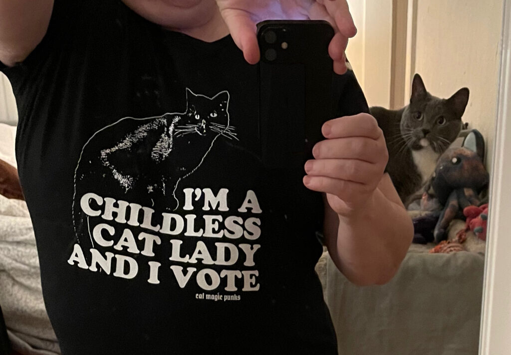 Photo of myself in the mirror, cropped to just the torso. I'm wearing a black T-shirt with a cat outline in white. Below that is white text: "I'M A CHILDLESS CAT LADY AND I VOTE." My gray and white cat "Eights" is photobombing me from her cat tree on the right side.