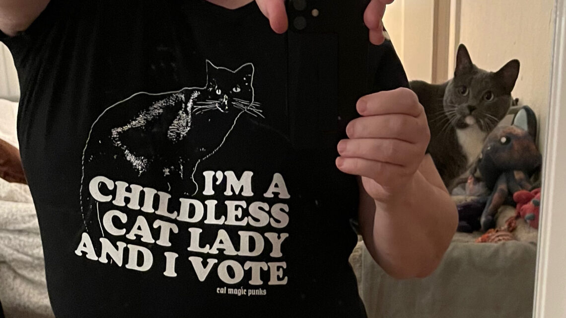 Photo of myself in the mirror, cropped to just the torso. I'm wearing a black T-shirt with a cat outline in white. Below that is white text: "I'M A CHILDLESS CAT LADY AND I VOTE." My gray and white cat "Eights" is photobombing me from her cat tree on the right side.